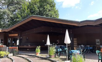 A cozy restaurant with a wooden facade and an inviting terrace. There are tables and chairs outside, surrounded by plants. | © Kulturcafe Riezlern | Veronika Senn