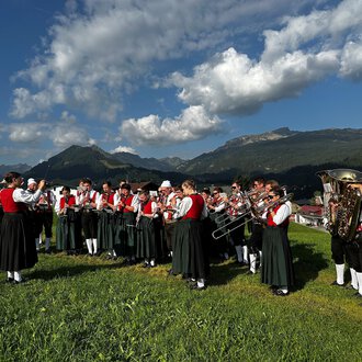 Eine Blaskapelle in traditioneller Tracht steht im Freien vor einem Holzhaus. Im Hintergrund sind grüne Wiesen und Berge zu sehen. | © Trachtenkapelle Riezlern