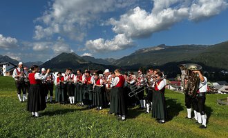 Eine Blaskapelle in traditioneller Tracht steht im Freien vor einem Holzhaus. Im Hintergrund sind grüne Wiesen und Berge zu sehen. | © Trachtenkapelle Riezlern