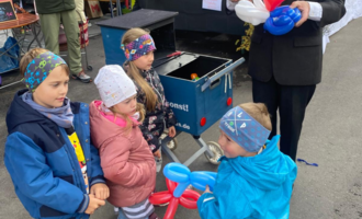 Four children are standing together and eagerly watching a man who is shaping balloons. They are wearing colorful clothing and are in a festive environment.