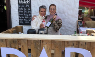 Two cheerful women are standing behind a wooden bar, holding drinks in their hands. In the background, a menu with various offers can be seen.