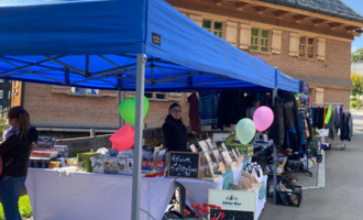 A market stall under blue tents with various goods. People are standing nearby and balloons are visible.