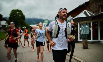 Eine Gruppe von Läufern joggt entlang einer Straße. Der Himmel ist bewölkt und die Umgebung wirkt bergig und grün. | © Kleinwalsertal Tourismus | Oliver Farys