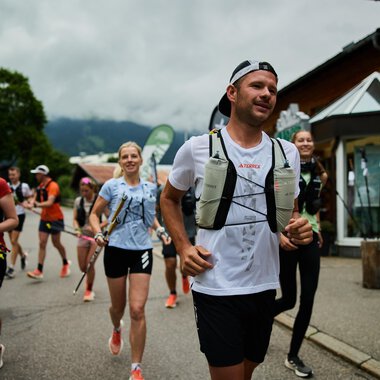 Eine Gruppe von Läufern joggt entlang einer Straße. Der Himmel ist bewölkt und die Umgebung wirkt bergig und grün. | © Kleinwalsertal Tourismus | Oliver Farys