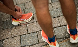 Ein Mann bindet seine Sportschuhe mit orangefarbener Sohle. Der Untergrund besteht aus grauen Pflastersteinen. | © Kleinwalsertal Tourismus | Oliver Farys