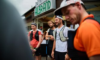 Eine Gruppe von Sportlern steht zusammen, während sie sich auf einen Wettkampf vorbereiten. Im Hintergrund ist ein Geschäftsschild zu sehen. | © Kleinwalsertal Tourismus | Oliver Farys