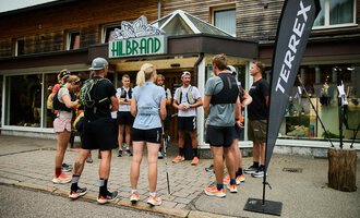 Eine Gruppe von Läufern versammelt sich vor einem Sportgeschäft. Im Hintergrund sind ein Schild und eine Werbeflagge zu sehen. | © Kleinwalsertal Tourismus | Oliver Farys
