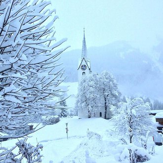 A snowy landscape with a church in the background. Trees and bushes are completely covered in snow.