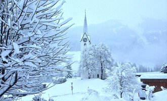A snowy landscape with a church in the background. Trees and bushes are completely covered in snow.