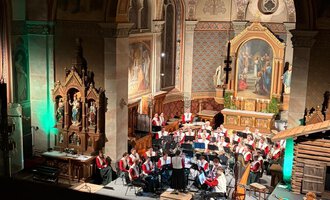 Eine große Musikkapelle in traditionellen Trachten steht auf einer Holzplattform mit Bergblick. Im Hintergrund sind wunderschöne Alpenlandschaften zu sehen. | © Trachtenkapelle Riezlern