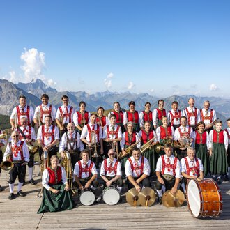 © Trachtenkapelle Riezlern Eine große Musikkapelle in traditionellen Trachten steht auf einer Holzplattform mit Bergblick. Im Hintergrund sind wunderschöne Alpenlandschaften zu sehen. | © Trachtenkapelle Riezlern