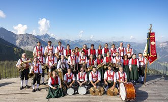 Eine große Musikkapelle in traditionellen Trachten steht auf einer Holzplattform mit Bergblick. Im Hintergrund sind wunderschöne Alpenlandschaften zu sehen. | © Trachtenkapelle Riezlern