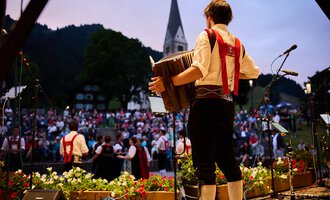 A musician plays an accordion in front of a large crowd. The atmosphere is festive, surrounded by a beautiful natural landscape. | © Kleinwalsertal Tourismus | Oliver Farys