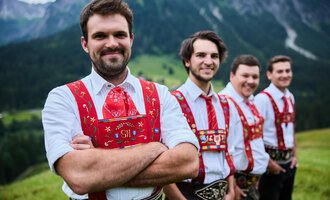 Four men in traditional attire stand in front of a picturesque mountain landscape. They smile kindly at the camera, the surroundings are green and hilly. | © Kleinwalsertal Tourismus | Oliver Farys