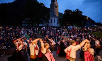 A festive dance performance takes place in front of a church. In the background, a large crowd can be seen watching. | © Kleinwalsertal Tourismus | Oliver Farys