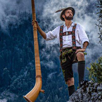 A man in traditional costume stands on a rock and holds an alphorn. In the background, mountains and fog can be seen.