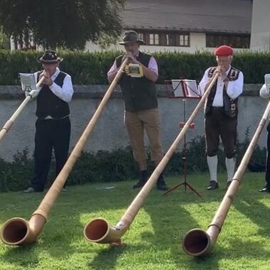 Fünf Männer in traditioneller Kleidung spielen auf langen Alphörnern im Freien. Die Szenerie ist von einer grünen Wiese und bayerischen Bergen umgeben. | © Alphornfreunde grenzenlos