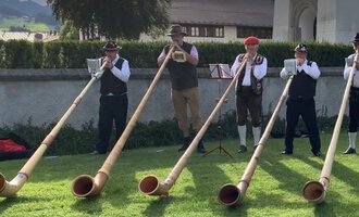 Eine Gruppe von fünf Männern spielt Alphörner auf einer Wiese. Sie tragen traditionelle Trachten und stehen in einer malerischen Umgebung. | © Alphornfreunde grenzenlos