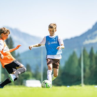 Junge Fußballspieler auf einem grünen Spielfeld mit Bergen im Hintergrund. Ein Spieler dribbelt den Ball, während ein anderer ihn versucht zu stoppen. | © Fußballschule Anstoß