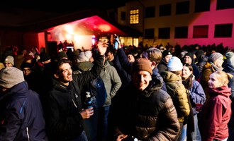 A lively crowd is celebrating at night under a tent-like pavilion. Many people are wearing warm clothing and seem to be having fun.