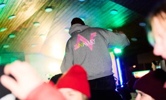 A person is standing on a platform wearing a gray hoodie with the inscription "Valley." In the background, colorful lights and the cheerful hustle and bustle of a celebration can be seen.