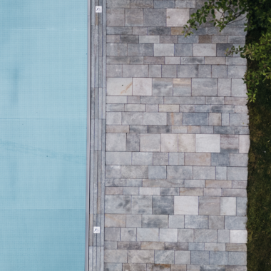 A modern designed outdoor area with a blue pool and a stone path. Next to it lies a green lawn and some foliage. | © Naturhotel Chesa Valisa