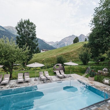 A beautiful pool with lounge chairs and umbrellas in a picturesque setting. In the background, green hills and mountains can be seen.