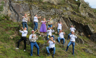 A group of musicians is standing on a green, grassy hill while playing various wind instruments. In the middle, one person is wearing a purple dress and is joyfully playing along.