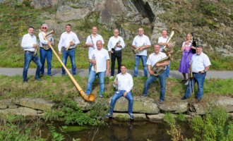 A group of musicians is standing outdoors, surrounded by nature. They are playing various instruments and wearing white shirts.