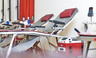 Ein Raum mit zwei Liegen, die für Blutspenden vorbereitet sind. Im Hintergrund sind weitere Ausstattungen und ein heller Raum mit roten Vorhängen zu sehen.