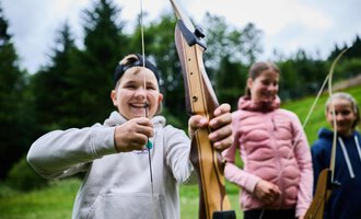 Zwei Personen im Wald beim Bogenschießen. Die Frau trägt einen bunten Pullover und zielt mit einem Bogen, während der Mann in einem grünen T-Shirt hinter ihr steht. | © Kleinwalsertal Tourismus eGen - Fotograf: Oliver Farys