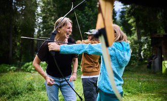 A woman stands in the forest aiming with a bow. Around her are lush green plants and trees. | © Kleinwalsertal Tourismus eGen - Fotograf: Oliver Farys