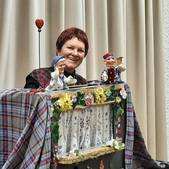 © Marie Luise Kaiser | Veronika Senn Eine Frau präsentiert ein Puppenspiel mit zwei handgefertigten Marionetten. Der Hintergrund ist mit einem hellen Vorhang und einer bunten Dekoration geschmückt. | © Marie Luise Kaiser | Veronika Senn