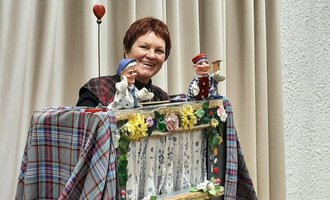 Eine Frau präsentiert ein Puppenspiel mit zwei handgefertigten Marionetten. Der Hintergrund ist mit einem hellen Vorhang und einer bunten Dekoration geschmückt. | © Marie Luise Kaiser | Veronika Senn