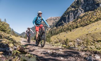 Zwei Radfahrer fahren auf einem Wanderweg durch die Berge. Die Landschaft ist grün und sonnig unter einem klaren blauen Himmel. | © Kleinwalsertal Tourismus | Bastian Morell