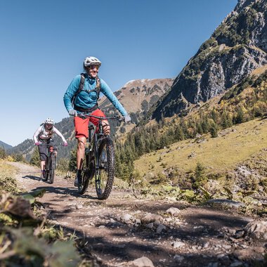 Zwei Radfahrer fahren auf einem Wanderweg durch die Berge. Die Landschaft ist grün und sonnig unter einem klaren blauen Himmel. | © Kleinwalsertal Tourismus | Bastian Morell