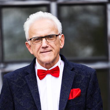 Ein älterer Mann mit grauen Haaren trägt einen eleganten Anzug und eine rote Fliege. Er hält ein Blatt Papier in der Hand und steht vor einem Gebäude mit großen Fenstern. | © Johann Mösenbichler - Fotograf: Tobias Epp