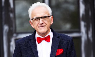 Ein älterer Mann mit grauen Haaren trägt einen eleganten Anzug und eine rote Fliege. Er hält ein Blatt Papier in der Hand und steht vor einem Gebäude mit großen Fenstern. | © Johann Mösenbichler - Fotograf: Tobias Epp