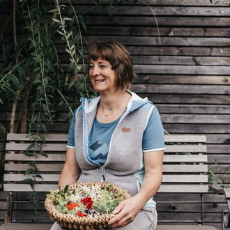 A smiling woman is sitting on a bench holding a basket of fresh plants. In the background, wooden slats and green plants can be seen. | © Naturhotel Chesa Valisa