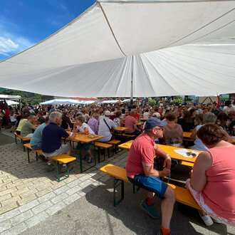 Eine große Menschenmenge sitzt an Tischen unter einem Sonnenschutz. Der Himmel ist blau und es herrscht eine festliche Atmosphäre.