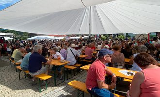 Eine große Menschenmenge sitzt an Tischen unter einem Sonnenschutz. Der Himmel ist blau und es herrscht eine festliche Atmosphäre.