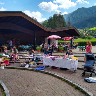 Ein lebhafter Flohmarkt im Freien mit vielen Tischen voller Waren. Im Hintergrund sind Stände, Kinder und eine malerische Berglandschaft zu sehen. | © @Flohmarkt im Kurpark Riezlern Kulturcafe