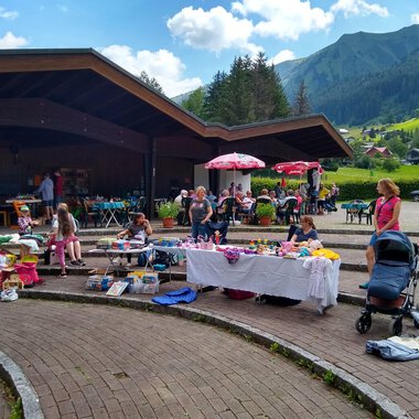 Ein lebhafter Flohmarkt im Freien mit vielen Tischen voller Waren. Im Hintergrund sind Stände, Kinder und eine malerische Berglandschaft zu sehen. | © @Flohmarkt im Kurpark Riezlern Kulturcafe