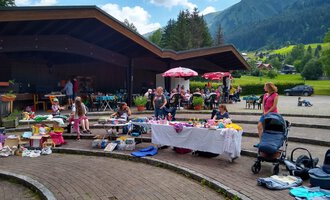 A lively outdoor flea market with many tables full of goods. In the background, there are stalls, children, and a picturesque mountain landscape. | © @Flohmarkt im Kurpark Riezlern Kulturcafe