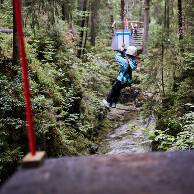 A child swings on a zip line between trees in a green forest. Surrounded by nature and a narrow stream below. | © Kleinwalsertal Tourismus | Oliver Farys
