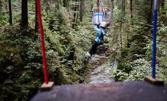 Ein Kind schwingt an einer Seilrutsche zwischen Bäumen in einem grünen Wald. Umgeben von Natur und einem schmalen Bach darunter. | © Kleinwalsertal Tourismus | Oliver Farys