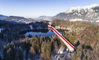 Eine lange Brücke mit roten und weißen Streifen führt über ein ruhiges, schneebedecktes Tal und einen See. Im Hintergrund sind majestätische Berge zu sehen, die in der Wintersonne erstrahlen.