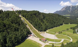 Eine beeindruckende Skisprungschanze umgeben von grünen Wäldern und Bergen. Die Landschaft ist malerisch und zeigt eine Mischung aus Natur und Sporteinrichtungen.
