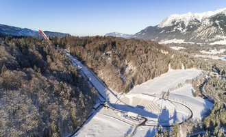 Eine schneebedeckte Landschaft mit Bäumen und einem klaren Himmel. Im Hintergrund sind Berge zu sehen, die die winterliche Szenerie ergänzen.