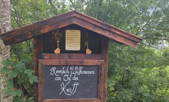A traditional wooden sign with the inscription "Warm Welcome to the Ötztal". Surrounded by green trees and a rural setting. | © Nicole Lughammer
